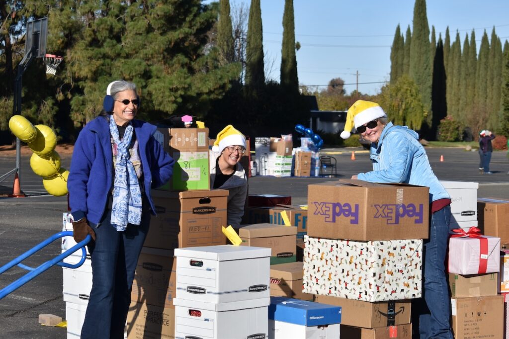 Three volunteers in festive hats and earmuffs smile while organizing holiday gift boxes outside